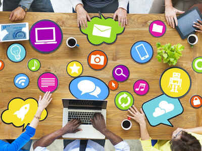 Mehrere Personen sitzen um einen Tisch herum, auf dem ein Laptop und verschiedene farbige Symbole für Technologie und Kommunikation liegen, wie z.B. ein Computer, ein Briefumschlag, eine Kamera, ein Smartphone und Wolken. Die Personen scheinen an einer Besprechung teilzunehmen und interagieren mit den Symbolen.