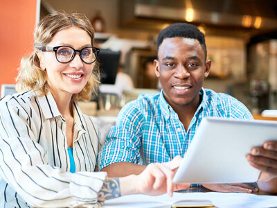 Zwei lächelnde Personen sitzen an einem Tisch und arbeiten gemeinsam. Die Frau links trägt eine Brille und ein gestreiftes Oberteil und zeigt auf ein Dokument oder ein Tablet, das vor dem Mann liegt. Der Mann rechts trägt ein kariertes Hemd und hält ein Tablet in der Hand. Beide wirken freundlich und konzentriert. Im Hintergrund sieht man eine gemütliche Umgebung, möglicherweise ein Café oder ein offener Arbeitsbereich. Die Atmosphäre wirkt kollegial und entspannt.