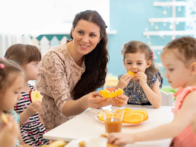 Eine Kindergruppe mit einer Erzieherin teilt Orangenscheiben an einem Tisch, während im Hintergrund eine Glas Orangensaft steht.