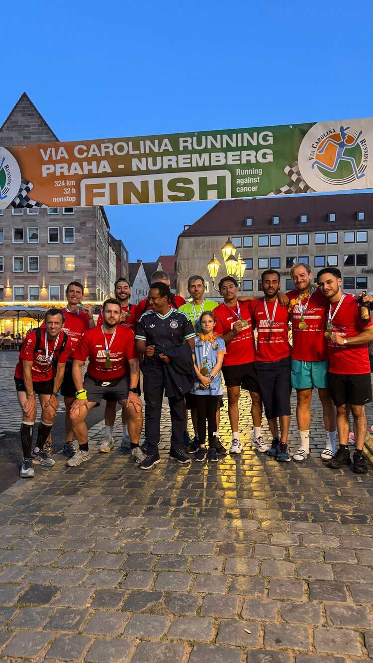 Gruppenfoto der Läuferinnen und Läufer unter dem Zielbanner des Via Carolina Running von Prag nach Nürnberg. Die Teilnehmenden tragen Medaillen und rote Trikots der ESO Education Group.