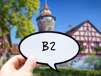 Eine Hand hält eine Sprechblase mit dem Text 'B2'. Im Hintergrund ist eine malerische Szene mit traditionellen europäischen Gebäuden zu sehen, darunter ein Fachwerkhaus und ein Gebäude mit einem turmartigen Aufbau. Im Hintergrund sind auch Bäume und Grünflächen sichtbar.