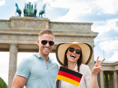 Zwei lächelnde Personen posieren vor dem Brandenburger Tor in Berlin. Die Person auf der linken Seite trägt eine Sonnenbrille und ein hellblaues Polohemd. Die Person auf der rechten Seite trägt einen großen Strohhut, eine Sonnenbrille und hält eine kleine deutsche Flagge. Beide machen ein Peace-Zeichen mit den Fingern. Im Hintergrund ist die Quadiga auf dem Brandenburger Tor zu sehen.