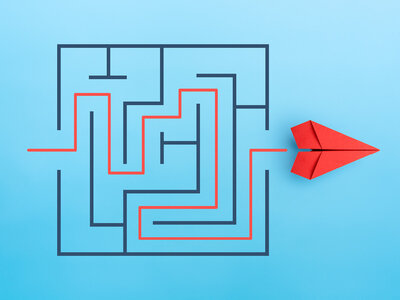 Ein Labyrinth mit einem roten Pfad, der den Weg durch das Labyrinth zeigt. Rechts neben dem Labyrinth liegt ein rotes Papierflugzeug. Das Labyrinth und das Flugzeug sind auf einem hellblauen Hintergrund platziert.