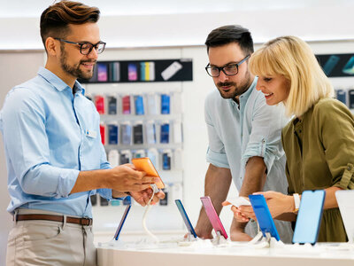Drei Personen stehen in einem Elektronikgeschäft und betrachten verschiedene Smartphones. Der Mann links, der ein hellblaues Hemd und eine Brille trägt, hält ein orangefarbenes Smartphone in der Hand und erklärt etwas. Das Paar rechts, ein Mann mit Brille und hellblauem Hemd sowie eine Frau mit blonden Haaren und grünem Oberteil, schauen interessiert auf die ausgestellten Smartphones in verschiedenen Farben wie Blau, Rosa und Schwarz. Im Hintergrund sind Regale mit weiteren Smartphone-Verpackungen zu sehen. Alle drei wirken freundlich und engagiert.