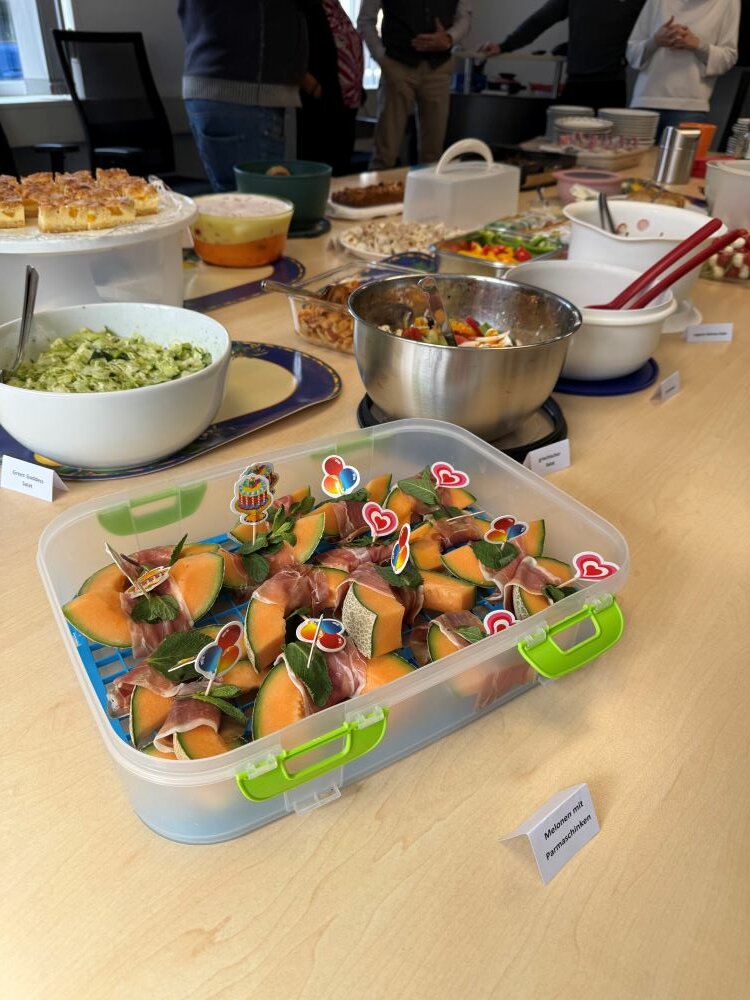 Achtsamtkeit im Fokus: ESO Education Group veranstaltet erneut erfolgreichen Gesundheitstag in Stockstadt Ein Tisch ist mit verschiedenen Snacks und Speisen gedeckt. Im Vordergrund befindet sich eine durchsichtige Box mit Melonenstücken, die mit Prosciutto und Minzblättern garniert sind.