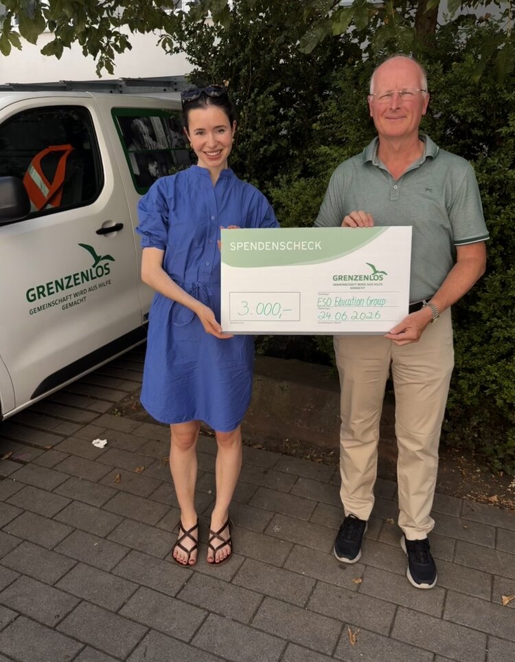 Eine Frauen in blauem Kleid und ein Mann in grauem Hemd lächeln in die Kamera und halten einen Spenden-Check der ESO Education Group hoch.