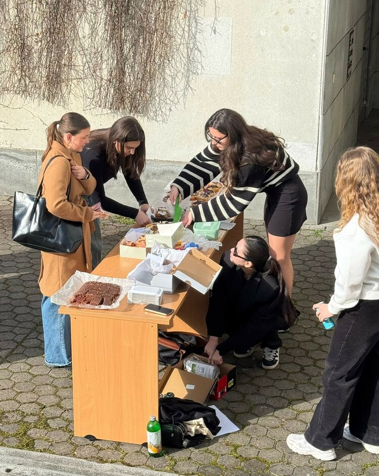 Global Money Week 2026 an der Euroskola Praha Mehrere Personen stehen oder knien um einen Tisch im Freien vor einem Gebäude, auf dem verschiedene Backwaren ausgelegt sind. Die Gruppe scheint sich an dem Stand zu bedienen.