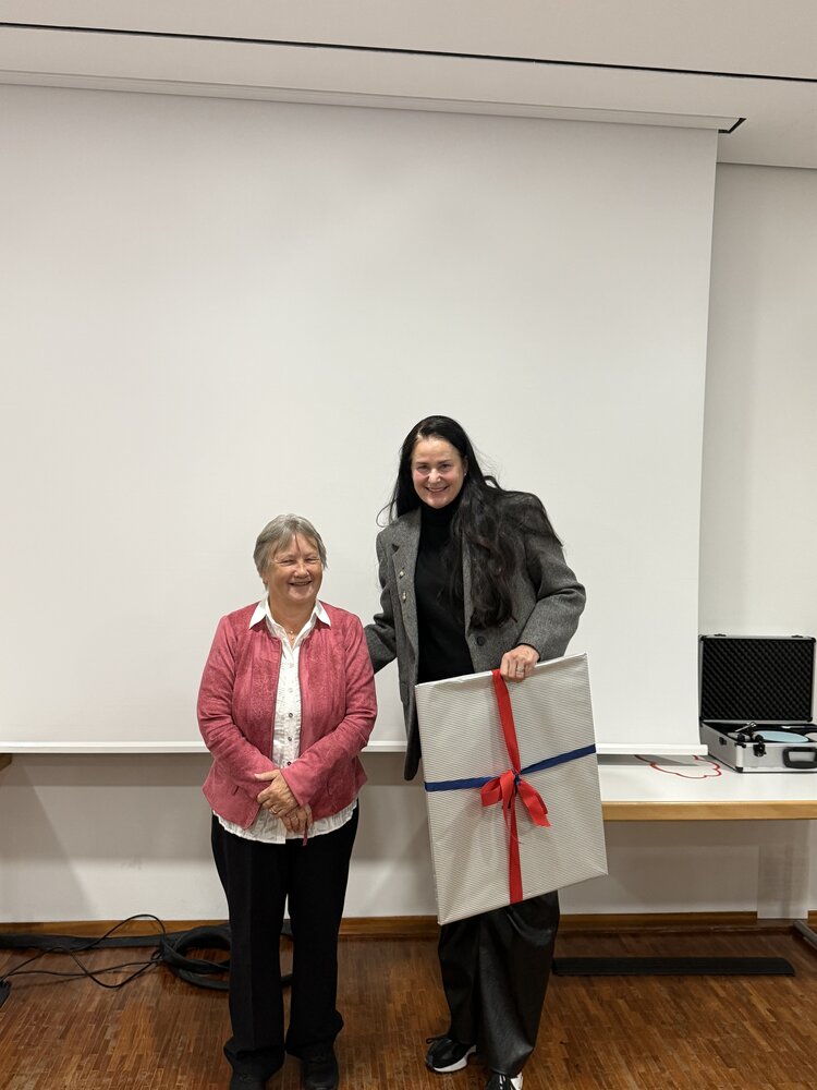 Zwei Frauen stehen in einem Seminarraum vor einer großen, weißen Projektionswand. Die kleinere Frau links trägt eine pinke Jacke über einer weißen Bluse und steht mit gefalteten Händen da. Die größere Frau rechts trägt einen grauen Blazer und schwarze Hose und hält ein großes, rechteckiges, eingepacktes Geschenk in der Hand.