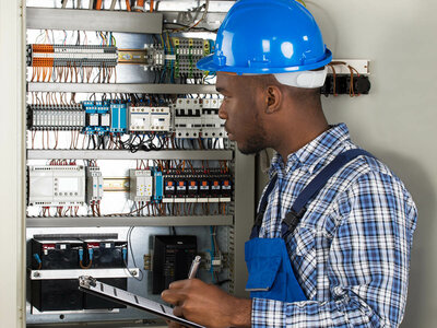 Ein Elektriker mit einem blauen Helm und einem karierten Hemd steht vor einem offenen Elektroschaltkasten. Er hält ein Klemmbrett und einen Stift in der Hand und scheint Notizen zu machen oder eine Inspektion durchzuführen. Der Schaltkasten ist mit verschiedenen Schaltern, Kabeln und Sicherungen ausgestattet. Der Elektriker trägt eine blaue Arbeitshose mit Werkzeugtaschen.