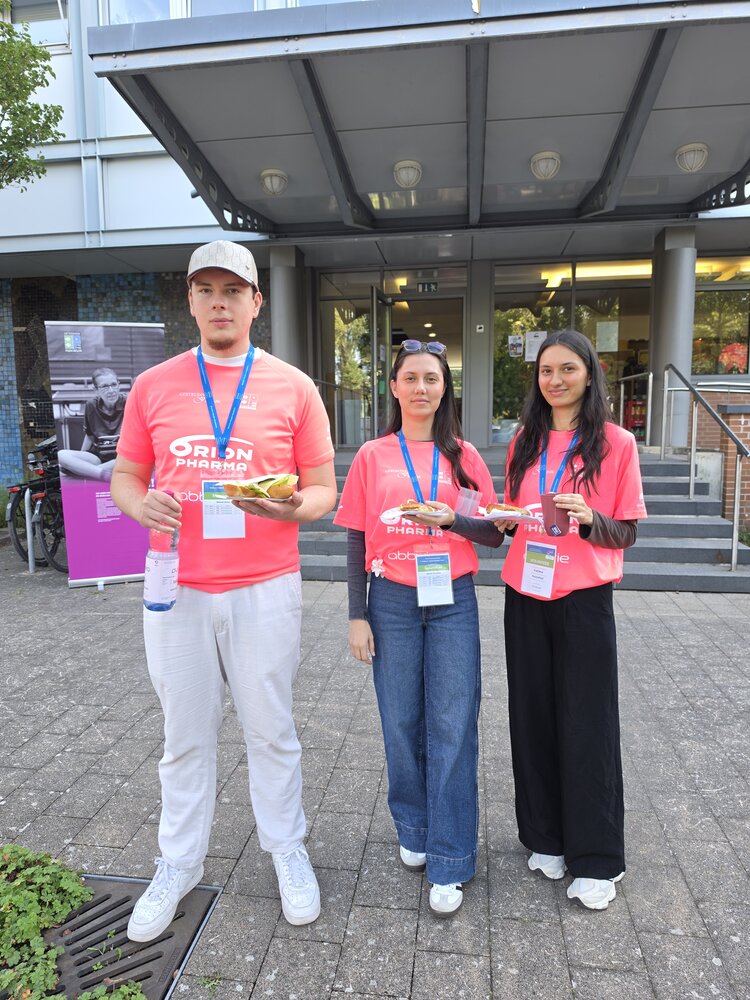 Gemeinsam im Einsatz für die PingPongParkinson-WM 2026 Vor einem Gebäude stehen drei Personen in pinken T-Shirts mit Ausweisbändern um den Hals. Die Personen halten Sandwiches und Getränke in den Händen.