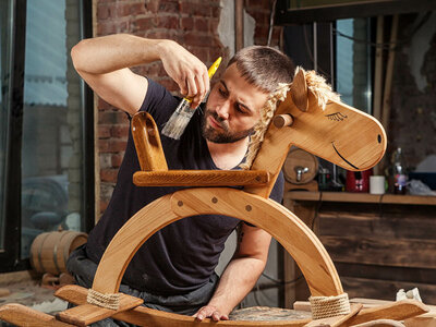 Ein Mann sitzt in einer Werkstatt und lackiert sorgfältig ein hölzernes Schaukelpferd. Er hält einen Pinsel in der Hand und konzentriert sich auf die Details des Pferdes, das eine Mähne aus Seil und ein lächelndes Gesicht hat. Im Hintergrund sind Werkzeuge und andere Holzarbeiten zu sehen.