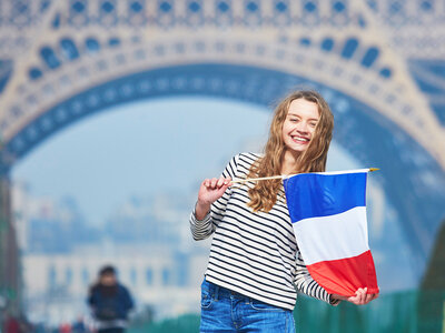 Eine lächelnde Frau steht vor dem Eiffelturm in Paris und hält eine französische Flagge in ihren Händen. Sie trägt ein gestreiftes Langarmshirt und Jeans. Im Hintergrund ist der Eiffelturm mit seiner charakteristischen Eisenkonstruktion zu sehen, und es scheint ein sonniger Tag zu sein.
