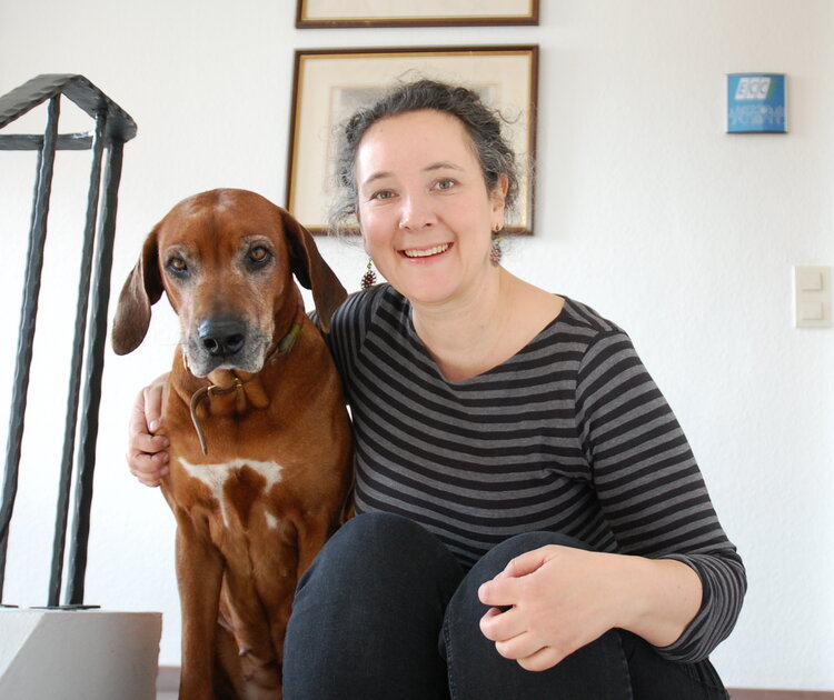 Mitarbeiterin Nadine Elbert, in schwarzer Jeans und schwarz-grau geringeltem Oberteil mit rötlichen Ohrringen, hat sich für das Foto auf die oberste Treppestufe gesetzt. Ihren rechten Arm hat sie um ihre Ridgebackhündin Cheina gelegt. Diese hat eine weiße Blesse auf der Brust und an Schnauze und um die Augen graue Haare und schaut interessiert in die Kamera. Im Hintergrund kann man ein Büroschild, auf dem "ECC" steht, erkennen.