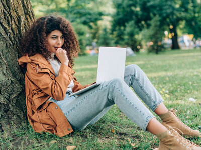 Eine Frau mit Locken, orangefarbener Jacke und Jeans sitzt auf einer Wiese an einen Baum gelehnt und schaut nachdenklich auf einen Laptop.