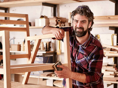 Eine Person in einer Werkstatt schleift einen Holzstuhlrahmen. Die Person trägt ein rot-schwarz kariertes Hemd und hält Schleifpapier, um das Holz zu glätten. Im Hintergrund sind Regale mit verschiedenen Werkzeugen und Materialien für die Holzbearbeitung zu sehen.