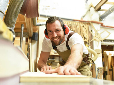 Ein Mann mit Gehörschutz arbeitet an einer Holzbearbeitungsmaschine in einer Werkstatt. Er trägt eine Schürze und ein weißes T-Shirt und scheint sich auf seine Arbeit zu konzentrieren. Im Hintergrund sind verschiedene Werkzeuge und Holzstücke zu sehen, die an der Wand und auf den Regalen angeordnet sind.