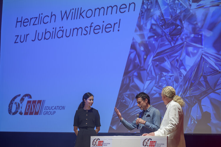Drei Personen auf der Bühne der Jubiläumsfeier der ESO Education Group; ein Mann spricht ins Mikrofon und gestikuliert.