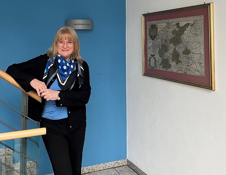 Mitarbeiterin steht lächelnd angelehnt an das Treppengeländer, trägt eine dunkle Hose, ein dunkles Jäckchen, ein Seidentuch in Blau mit weißen Punkten.