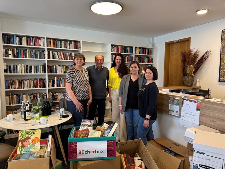 Silvia Semidei, CEO der ESO Education Group, posiert gemeinsam mit Sigrid Kaiser-Becker und einer Kollegin vom Logophon Verlag und Harry Kimmich, dem Vorsitzenden des Vereins Grenzenlos e.V., für ein Gruppenfoto. Im Hintergrund stehen weiße Bücherregale voller Bücher, links ein kleines, rundes Tischchen, auf dem eine große Kanne Kaffee, Tassen und eine Flasche Wasser stehen. Im Vordergrund stehen mehrere geöffnete Kartons, aus denen die zahlreichen gespendeten Bücher herausschauen. 