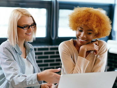 Zwei Personen sitzen an einem Tisch und arbeiten zusammen an einem Laptop. Eine Person hat kurze blonde Haare und trägt eine Brille, die andere hat voluminöse, lockige Haare und trägt einen beigen Pullover. Beide lächeln und wirken konzentriert.