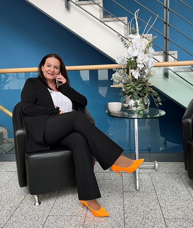 Mitarbeiterin Margot Niklas sitzt auf schwarzem Sessel im Treppenhaus, neben ihr ein Glastisch mit Vase und weißen Schnittblumen.