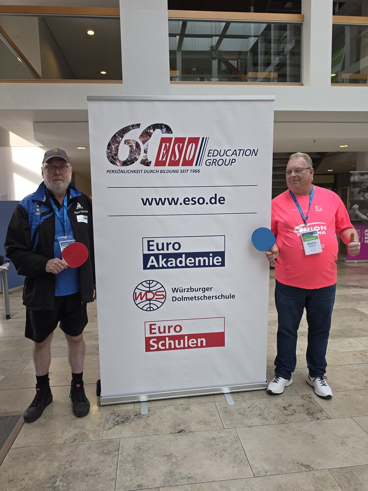 Gemeinsam im Einsatz für die PingPongParkinson-WM 2026 Zwei Männer mit Tischtennisschlägern stehen links und rechts neben einem Banner der ESO Education Group in einem Innenbereich.
