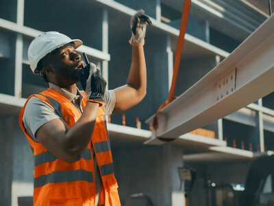 Ein Bauarbeiter trägt eine orange Warnweste, einen weißen Schutzhelm und Handschuhe. Er steht auf einer Baustelle und hält mit einer Hand einen Metallträger fest, während er mit der anderen Hand ein Funkgerät ans Ohr hält, um zu kommunizieren. Der Bauarbeiter wirkt konzentriert und engagiert. Im Hintergrund sind Teile eines Gebäudes oder einer Konstruktion zu sehen, die sich im Bau befindet. Die Szene vermittelt einen Eindruck von Teamarbeit und Sicherheit auf der Baustelle.