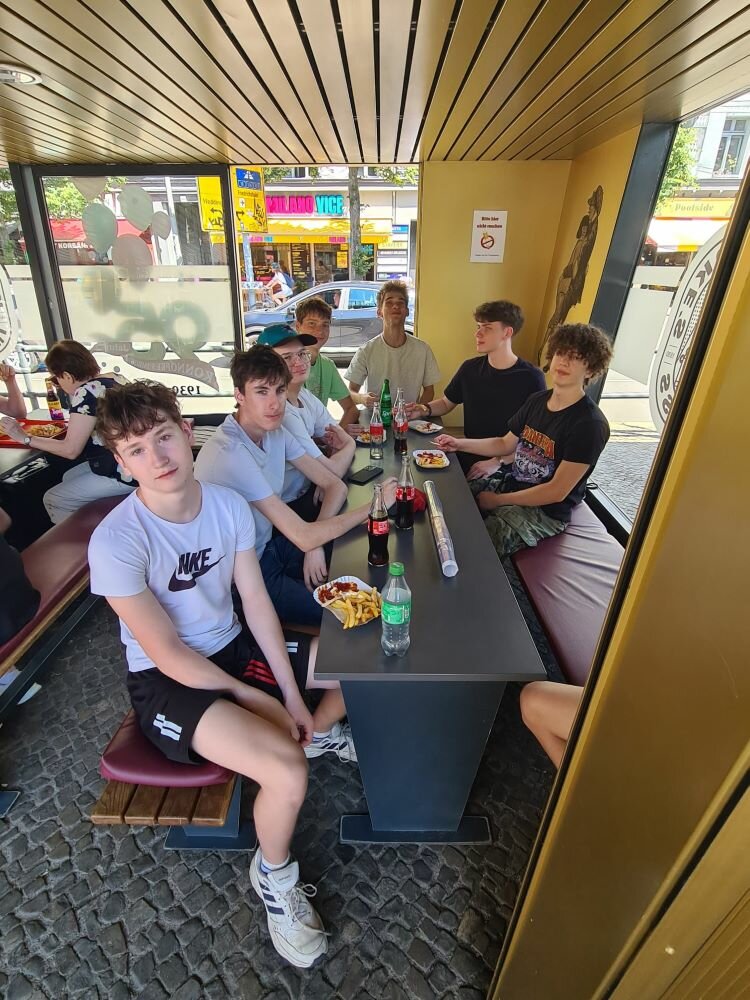 Klassenfahrt der Euroskola Prag nach Berlin Eine Gruppe von Jugendlichen sitzt an einem langen Tisch in einem Café mit gelben Wänden und großen Fenstern. Auf dem Tisch stehen Getränke, Snacks und persönliche Gegenstände wie Handys.