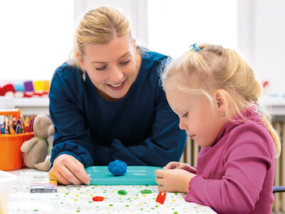 Eine Frau und ein kleines Mädchen sitzen an einem Tisch und basteln gemeinsam. Die Frau trägt ein dunkles Oberteil und lächelt freundlich, während sie dem Mädchen hilft. Das Mädchen, mit blonden Haaren und einem rosa Pullover, konzentriert sich auf das Bastelprojekt vor ihr. Auf dem Tisch liegen bunte Bastelmaterialien wie Knetmasse, Schere, Klebestift und verschiedene Papiere. Im Hintergrund sind ein Behälter mit Buntstiften und ein kleiner Stofftier-Hase zu sehen. Beide wirken glücklich und engagiert. Die Szene vermittelt eine warme und kreative Atmosphäre.