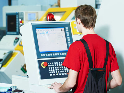 Eine Person in einem roten Hemd und schwarzen Overalls bedient ein großes Bedienfeld mit verschiedenen Knöpfen, Schaltern und einem Bildschirm, der eine Benutzeroberfläche anzeigt. Im Hintergrund sind weitere Maschinen und Geräte zu sehen.
