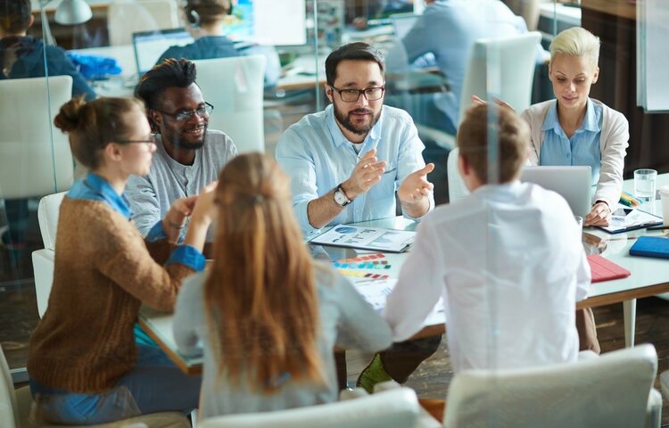 Eine Gruppe junger Berufstätiger sitzt in einem modernen Konferenzraum um einen Tisch und diskutiert lebhaft. Auf dem Tisch liegen Laptops, Notizbücher und Diagramme. Die Atmosphäre wirkt engagiert und kollaborativ, während ein Mann in der Mitte gestikulierend spricht. Alle Beteiligten wirken konzentriert und in das Gespräch vertieft.