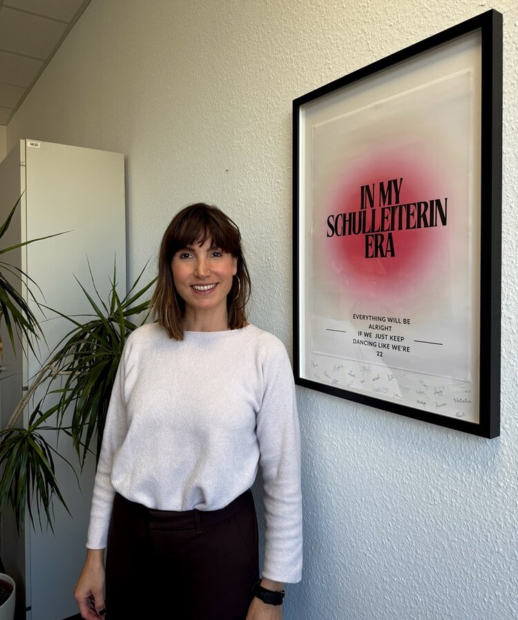 Simone Scharrer, Leitung der WDS, posiert lächelnd und an die rauhfasertapezierte gelehnt für ein Foto. Sie trägt eine dunkle Hose und ein weißes Langarmoberteil sowie braune Haare im Longbob mit Pony. 