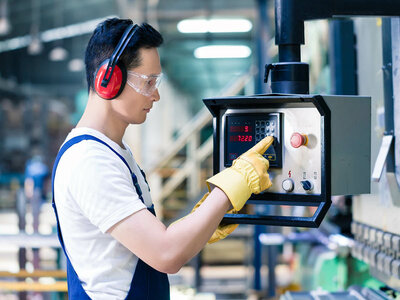 Ein Mann mit einem weißen T-Shirt und einer blauen Schürze steht in einer industriellen Umgebung und bedient das Bedienfeld einer Maschine. Er trägt gelbe Arbeitshandschuhe, eine Schutzbrille und einen Gehörschutz. Die Maschine hat ein digitales Display und verschiedene Bedienelemente. Im Hintergrund sind weitere Maschinen und Geräte zu sehen, die auf eine industrielle Umgebung hinweisen.