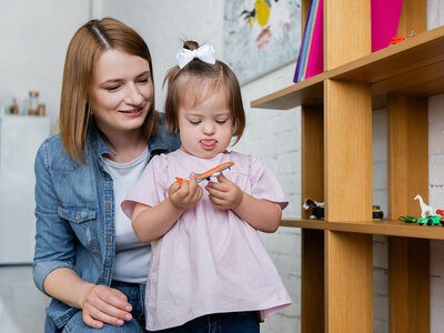 Eine Frau und ein Kind stehen in einem hellen, kindgerecht eingerichteten Raum. Die Frau, die eine blaue Denim-Jacke und ein weißes T-Shirt trägt, beugt sich leicht vor und unterstützt das Kind liebevoll. Das Kind trägt ein hellrosa Kleid und eine weiße Schleife im Haar. Es hält ein orangefarbenes Spielzeug in den Händen und blickt konzentriert darauf. Im Hintergrund ist ein hölzernes Regal mit Spielzeug und Büchern zu sehen, darunter bunte Bauklötze und eine weiße Zielfigur. Die Atmosphäre wirkt warm, fürsorglich und spielerisch.