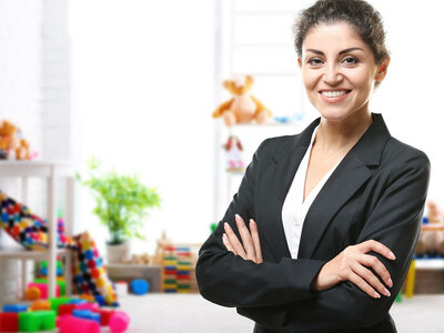Auf dem Bild ist eine Frau im Anzug zu sehen. Im Hintergrund befinden sich bunte Spielzeuge, darunter Bausteine, Stofftiere und Pflanzen. Die Szenerie wirkt lebhaft und farbenfroh.