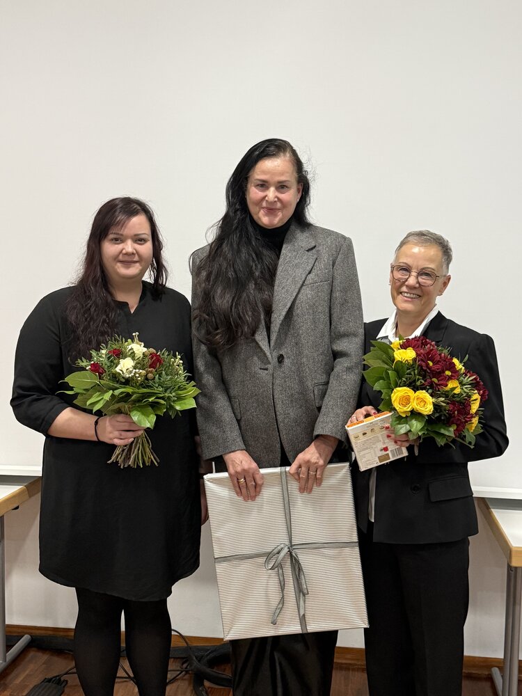 Drei Frauen stehen nebeneinander vor einer weißen Wand und schauen lächelnd in die Kamera; die Frau in der Mitte trägt einen grauen Blazer und hält ein großes, hell verpacktes Geschenk, während die Personen links und rechts in dunkler Kleidung jeweils einen farbigen Blumenstrauß in den Händen halten.