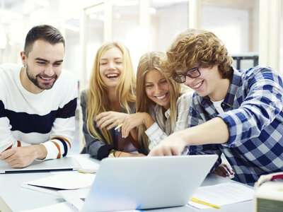 Vier junge Erwachsene sitzen an einem Tisch und arbeiten gemeinsam an einem Laptop. Die Person links, ein Mann mit Bart und einem gestreiften Pullover, lächelt. Die beiden Frauen in der Mitte, mit langen blonden Haaren, scheinen sich über etwas auf dem Laptop zu freuen. Der Mann rechts, mit Brille und einem karierten Hemd, zeigt auf den Laptop-Bildschirm. Auf dem Tisch liegen verschiedene Papiere und ein Stift.