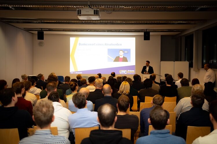 Großer Vortragsraum beim 25-jährigen Jubiläum des IT-Centers Dortmund: Zahlreiche Besucherinnen und Besucher sitzen auf Holzstühlen und verfolgen eine Präsentation auf einer großen Leinwand. Auf der Bühne steht ein Redner im Anzug am Podium und spricht zum Publikum. Die Leinwand zeigt eine Folie mit dem Titel „Dankeswort eines Absolventen“. Der Raum ist modern beleuchtet, an der Decke hängen technische Installationen und ein Beamer. Die Stimmung ist konzentriert und feierlich.