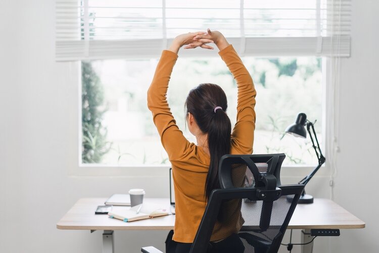 In einem hellen Büro ist eine Frau von hinten an einem Schreibtisch am Fenster zu sehen. Die Frau streckt die Arme mit verschränkten Fingern hoch über den Kopf und dehnt sich.