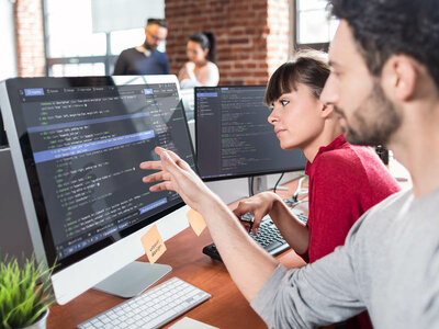 Eine Gruppe von Menschen arbeitet in einem Büro. Es sind zwei Computermonitore sichtbar, auf denen Code angezeigt wird. Eine Person zeigt auf den Bildschirm des linken Monitors, der mit einem Haftnotiz versehen ist, auf dem 'Nicht berühren' steht. Im Hintergrund sind weitere Personen und Büromöbel zu sehen.