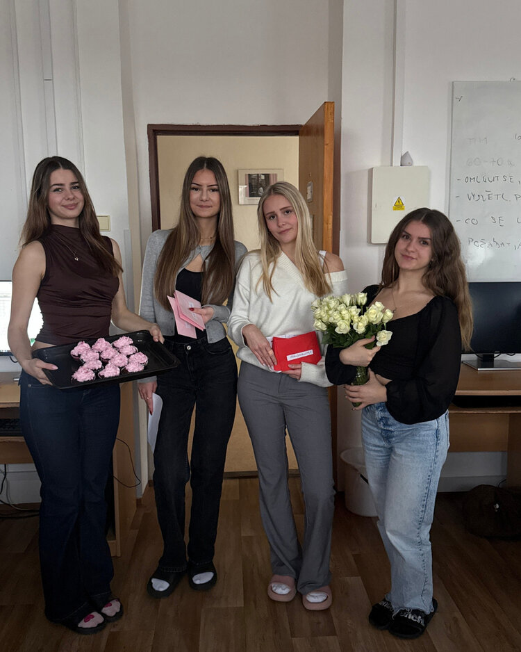 Vier junge Frauen stehen nebeneinander in einem Raum. Die erste Frau hält ein Tablett mit rosa verzierten Cupcakes, die zweite und dritte halten jeweils eine Karte und ein kleines Geschenk, während die vierte Frau einen Strauß weißer Rosen trägt. Alle lächeln in die Kamera.