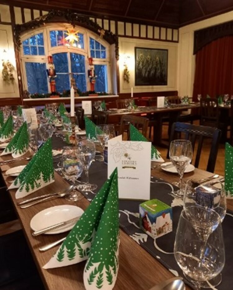 Lang eingedeckter Festtisch im Jugendstilsaal mit grünen Tannenbaum-Servietten, Menükarten, Gläsern und Kerzen, dekoriert für eine Weihnachtsfeier.