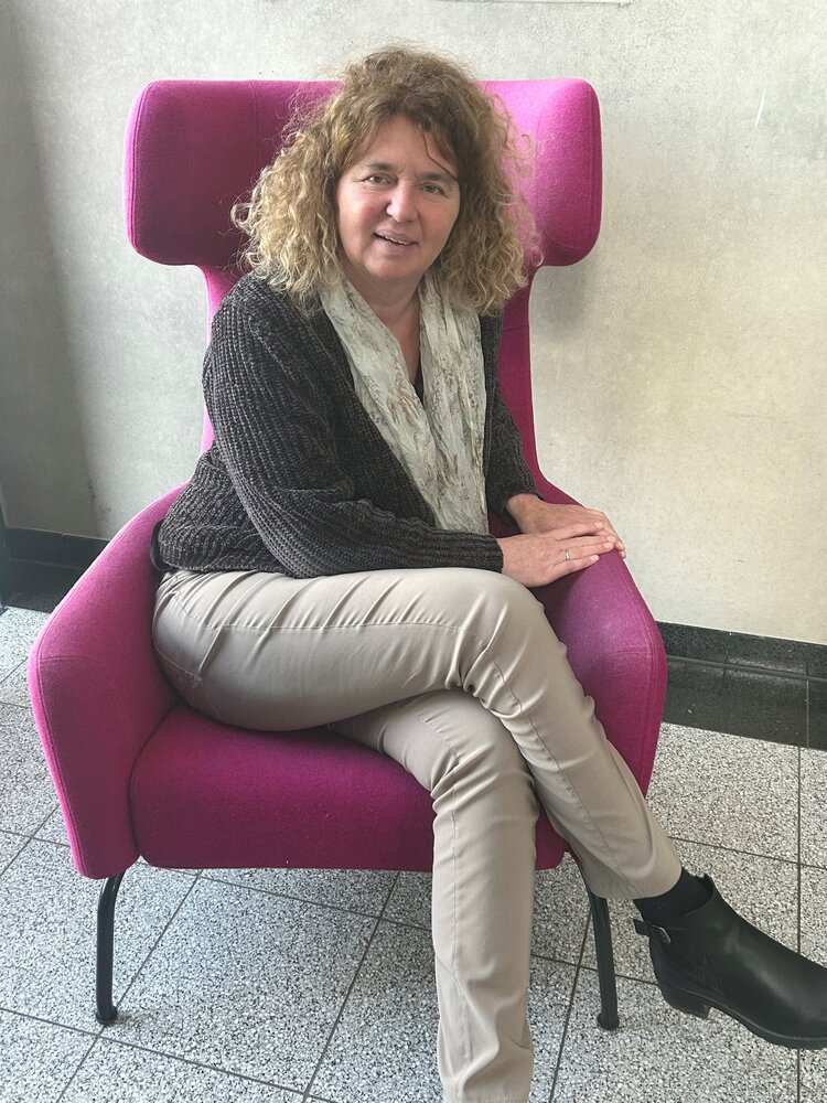 Die Mitarbeiterin Brigitte Berberich sitzt mit übereinandergeschlagenen Beinen in einem magentafarbenen Sessel in einem Flur oder einer Hotellobby. Sie hat dunkelblonde Locken und träg eine beigefarbene Hose mit einem dunklen Strickpullover und einem hellen Halstuch. Ihre Hände hat sie auf die linke Sessellehne gelegt.