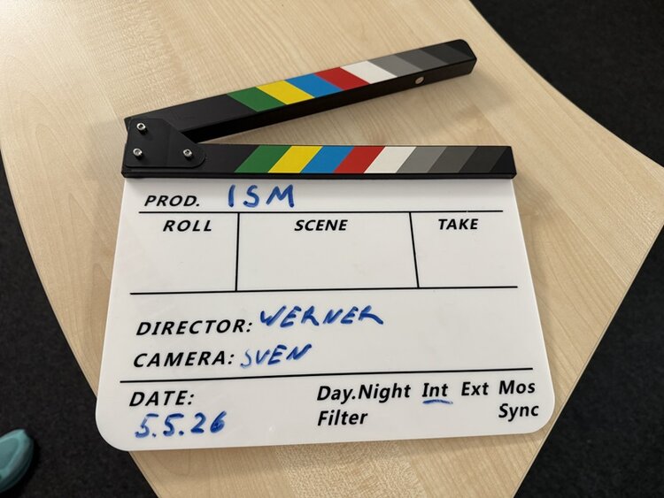 Eine Filmklappe mit farbigen Streifen am oberen Rand liegt auf einer hellen Holzoberfläche. Die Klappe ist geöffnet. Auf der weißen Fläche darunter sind handschriftliche Einträge in blauer Farbe zu sehen, darunter die Begriffe 'DIRECTOR: WERNER', 'CAMERA: SVEN' und das Datum '5.5.26'. Weitere vorgedruckte Felder wie 'PROD.', 'ROLL', 'SCENE', 'TAKE', 'Day.Night', 'Int', 'Ext', 'Filter', 'Mos', 'Sync' sind sichtbar, aber größtenteils nicht ausgefüllt. Ein Teil eines türkisfarbenen Schuhs ist am unteren Bildrand zu erkennen.