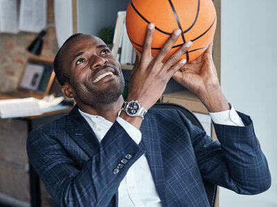 Ein Mann in einem Anzug hält einen Basketball in der Hand und bereitet sich darauf vor, ihn zu werfen. Er trägt eine Armbanduhr und steht in einem Büro mit Regalen und Papieren im Hintergrund. Der Mann lächelt und scheint sich auf den Wurf zu konzentrieren.