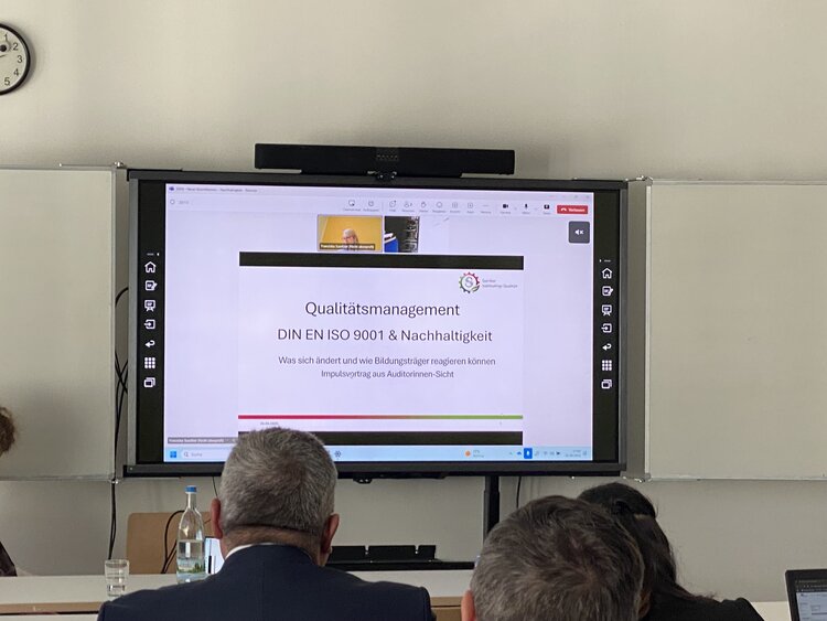 Eine Präsentation zum Thema „Qualitätsmanagement – DIN EN ISO 9001 & Nachhaltigkeit“ wird auf einem großen Bildschirm gezeigt. Mehrere Personen sitzen im Seminarraum und folgen dem Vortrag aufmerksam.