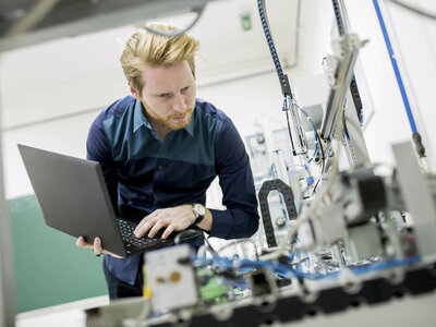 Eine Person arbeitet mit einem Laptop in einer industriellen oder laborähnlichen Umgebung. Um sie herum befinden sich verschiedene Maschinen und Geräte, möglicherweise im Bereich Automatisierung oder Robotik. Das Gesicht der Person ist aus Datenschutzgründen unscharf.