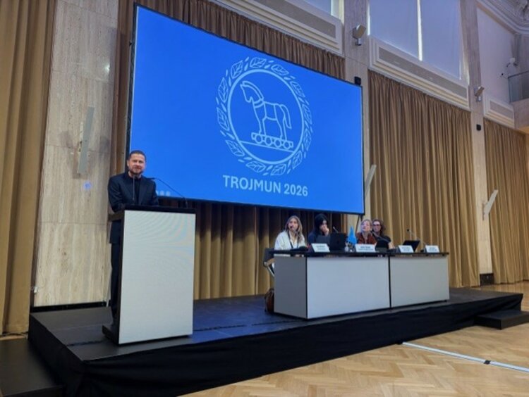 Ein Mann in einem dunkelblauen Anzug spricht an einem Rednerpult auf einer Bühne. Im Hintergrund ist eine Leinwand mit dem Logo und dem Schriftzug „TROJMUN 2026“ zu sehen. Vier Personen sitzen an einem Tisch rechts auf der Bühne.