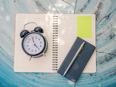 Ein offenes Notizbuch liegt auf einer blauen, strukturierten Oberfläche. Auf der linken Seite des Notizbuchs steht ein klassischer Wecker, der 10:10 Uhr anzeigt. Auf der rechten Seite des Notizbuchs liegt ein schwarzes Etui mit einem Stift darin. Ein gelber Haftnotizzettel ist oben rechts auf der Seite befestigt.
