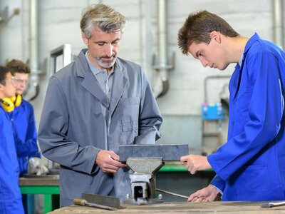 Drei Personen befinden sich in einer Werkstatt. Zwei von ihnen stehen an einer Werkbank und untersuchen ein Metallstück, das in einem Schraubstock eingespannt ist. Sie tragen blaue Laborkittel, eine Person hat eine Schutzbrille um den Hals. Im Hintergrund sind verschiedene Werkzeuge und Geräte zu sehen, wie sie typischerweise in einer Werkstatt vorkommen.